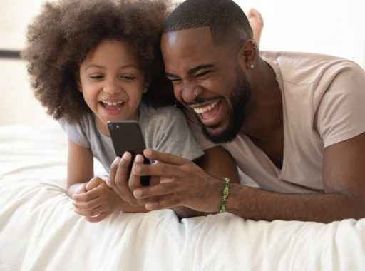Uma criança e estudante secundarista com o seu pai. Ambos estão felizes olhando para o Joov, o melhor comparador de planos de saúde, no celular.